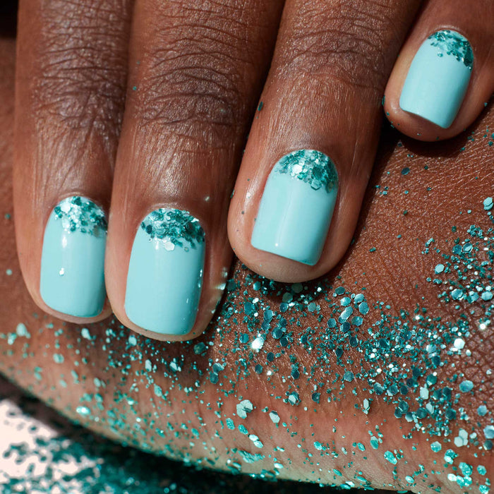 A close-up of a hand with light blue polish, featuring Manucurist Underwater Glitter—turquoise biodegradable accents by the cuticles applied with a nail art brush. Some glitter is also scattered on the skin beneath the hand.
