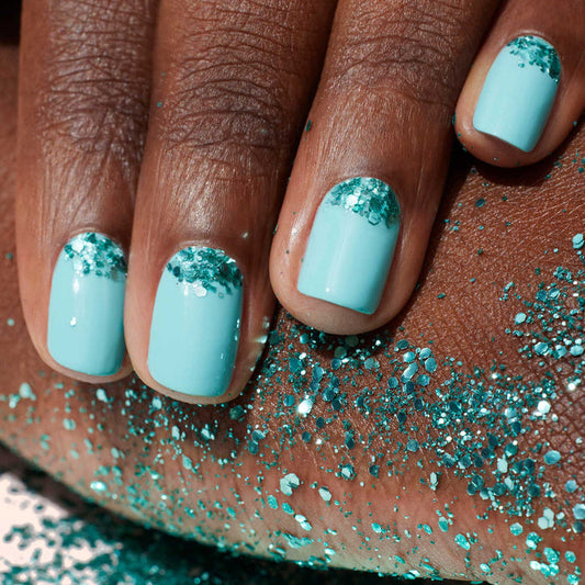 A close-up of a hand with light blue polish, featuring Manucurist Underwater Glitter—turquoise biodegradable accents by the cuticles applied with a nail art brush. Some glitter is also scattered on the skin beneath the hand.