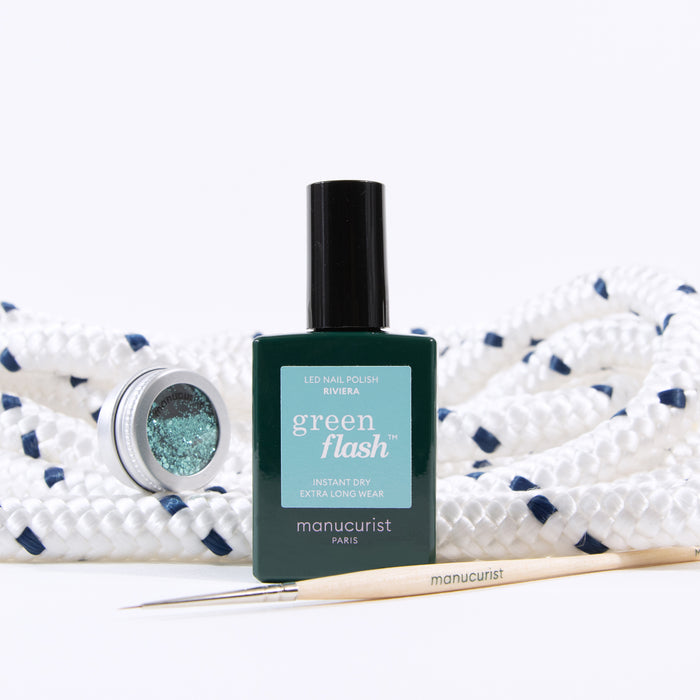 A bottle of Manucurist Underwater Glitter nail polish, a small open jar of biodegradable glitter, and a wooden manicure tool are arranged on a coiled white rope with navy accents.