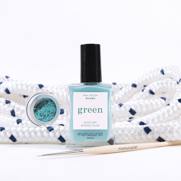 A bottle of Manucurist Underwater Glitter nail polish sits before coiled white rope with navy accents, joined by a small pot of biodegradable glitter and a fine nail art brush for creative designs.