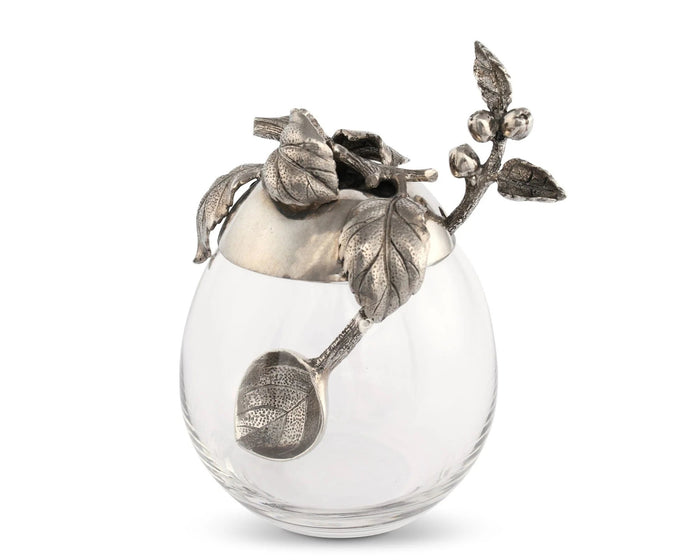 The VAGABOND HOUSE - APPLE HONEY POT features a decorative glass jar with a pewter cap showcasing intricate leaf and berry designs. The cap's handle mimics a stem with leaves, adding ornate elegance to this centerpiece by VAGABOND HOUSE, ideal for outdoor dining.
