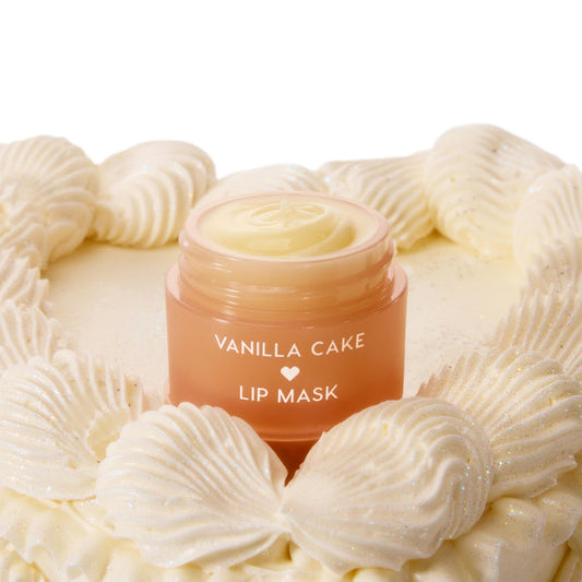 Vanilla Cake ColourPop Lip Mask jar, lid off to reveal creamy pale-yellow balm, set atop a vanilla cake with glittery piped frosting; peach ombre container with white lettering and a heart icon.