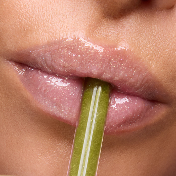 Close-up glossy lips wearing Colourpop Fresh Kiss Glossy Balm Vanilla Matcha shade, holding a shimmery green lip applicator between them, promoting hydrating Lip Gloss & Lip Oil finish