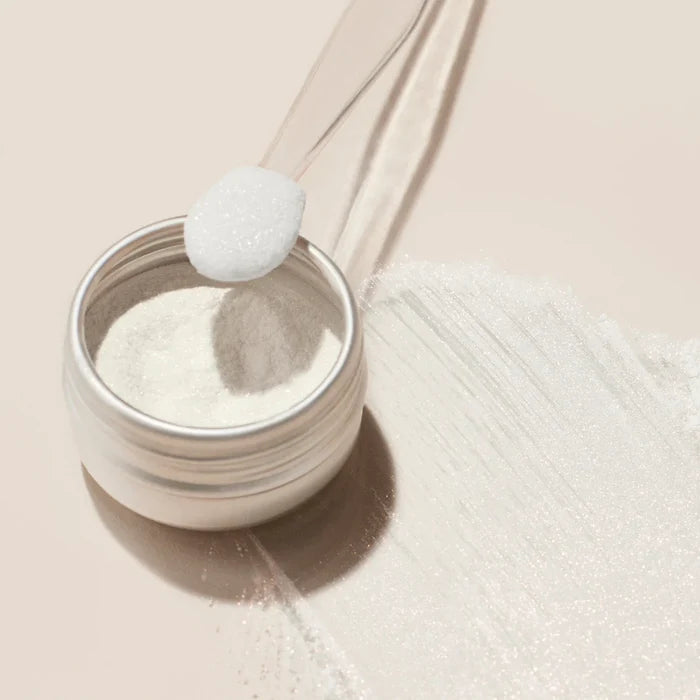 A small jar of Manucurist White Glazed Effect Powder sits open on a light surface, with a clear spoon holding a scoop above it and a streak of powder beside—ideal for creating stunning white glazed nails in your next manicure.
