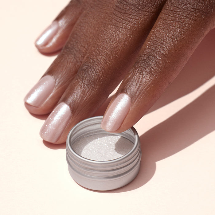 A hand with light pink, shimmery nails featuring a subtle white glazed effect created by Manucurist's White Glazed Effect Powder rests beside an open tin of the same powder on a pale surface.