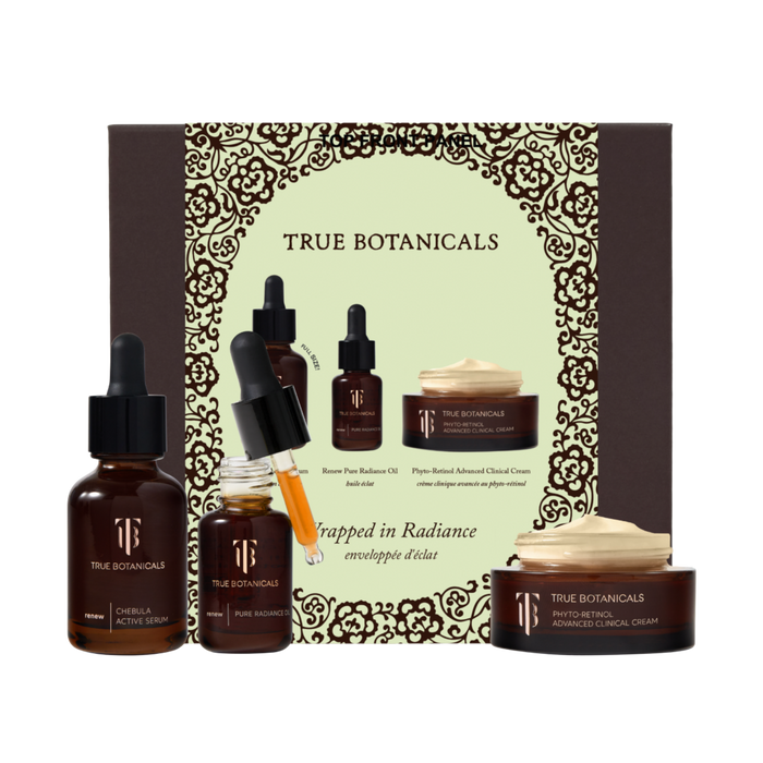 A product display of the True Botanicals "Wrapped in Radiance" gift set against a white background. In the center sits a large, dark brown box featuring an intricate golden filigree border framing the brand name. Arranged in front of the packaging are three skincare items: an amber glass bottle of Chebula Active Serum on the left, an open bottle of Renew Pure Radiance Oil with a yellow-tinted liquid dripping from its glass dropper in the middle, and a low, round jar of Phyto-Retinol Advanced Clinical Cream