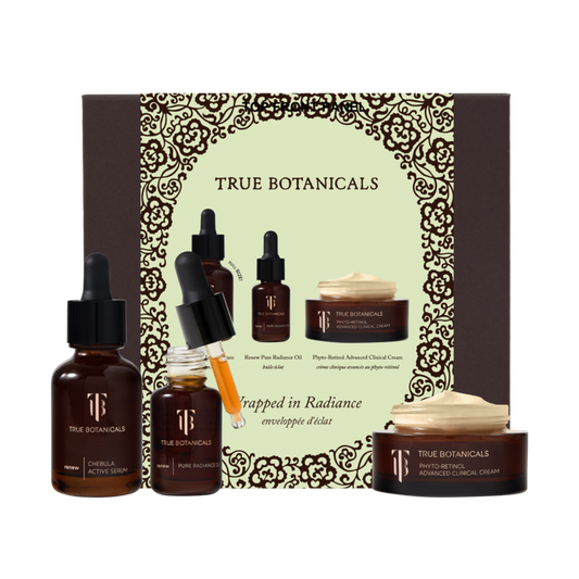 A product display of the True Botanicals "Wrapped in Radiance" gift set against a white background. In the center sits a large, dark brown box featuring an intricate golden filigree border framing the brand name. Arranged in front of the packaging are three skincare items: an amber glass bottle of Chebula Active Serum on the left, an open bottle of Renew Pure Radiance Oil with a yellow-tinted liquid dripping from its glass dropper in the middle, and a low, round jar of Phyto-Retinol Advanced Clinical Cream